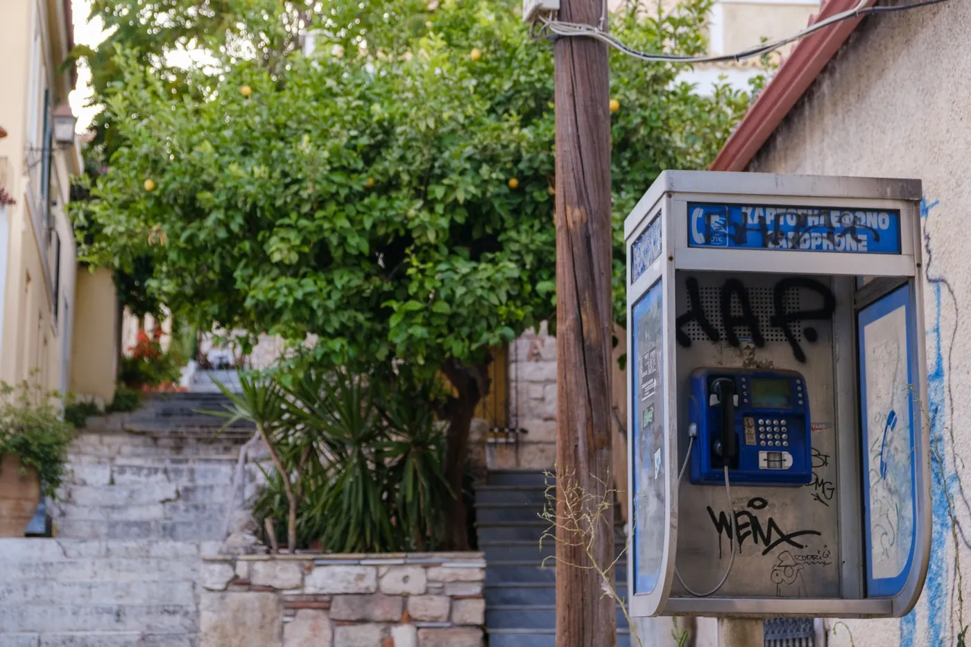 עמדת טלפון ציבורי כחולה עם גרפיטי ברחוב ביוון, לצד עץ הדר וקיר אבן, באור יום