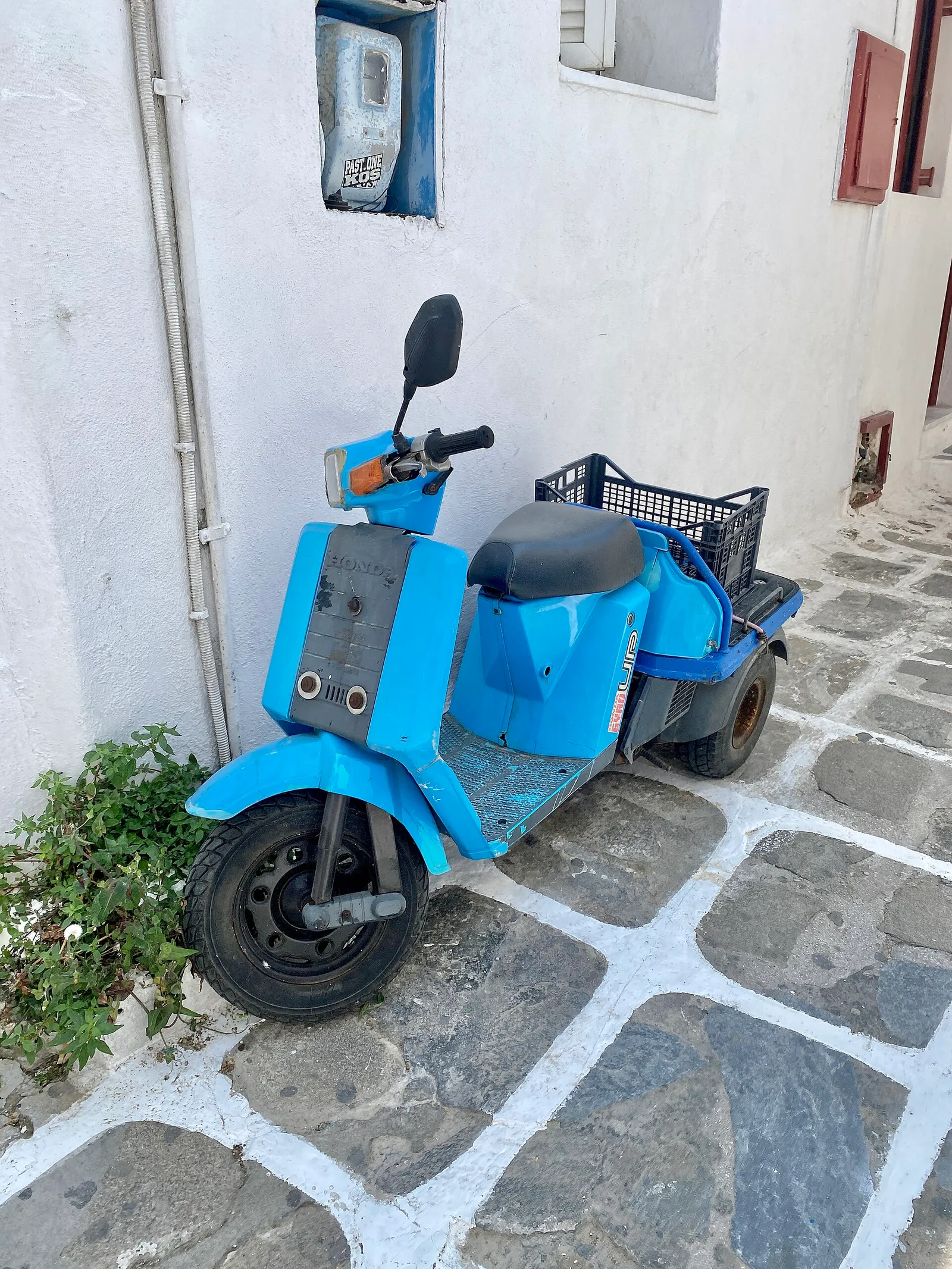 קטנוע כחול חונה בסמטה מרוצפת ולבנה במיקונוס ביום בהיר, כלי הרכב הפופולרי באיים שדורש הרחבת ביטוח