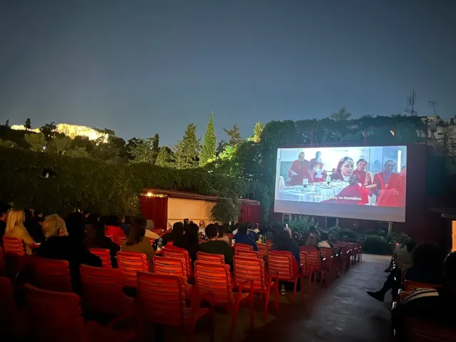 קולנוע פתוח בלילה עם כיסאות אדומים, קהל צופה ומסך מואר בין העצים