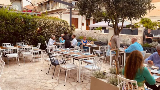 חצר מרוצפת עם כיסאות לבנים, עץ זית נטוע וסועדים בשולחנות