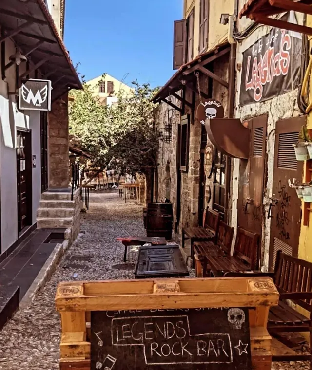 סמטה מרוצפת חלוקים עם ספסלי עץ, שלטים צבועים ולוח גיר בכניסה