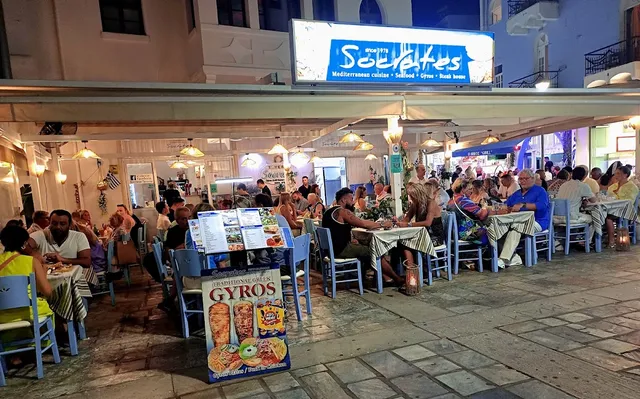 טברנה הומה בערב עם שולחנות על המדרכה, מפות משובצות ושלט מואר מעל