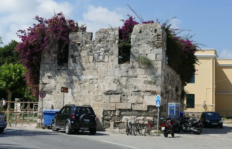 שריד חומת אבן מימי הביניים מכוסה בוגנוויליה בעיר העתיקה של קוס, עם רחוב ומכוניות חונות בחזית