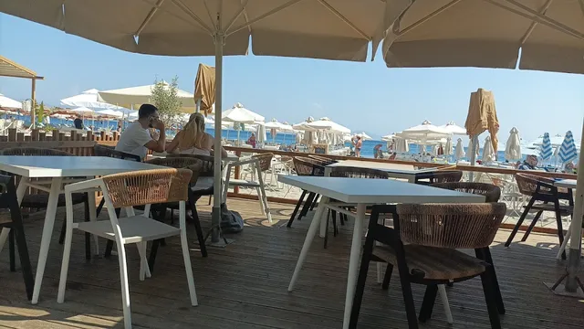 שולחנות לבנים וכיסאות קלועים על דק עץ תחת שמשיות, מול חוף הומה
