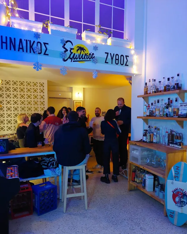בר קטן ומואר בערב, אנשים עומדים עם בירות ומדפי בקבוקים לצד קיר