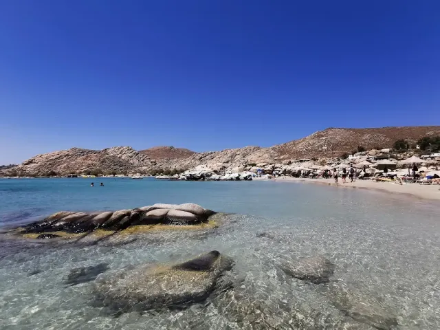 מים רדודים וצלולים עם סלעים מעוגלים, ומאחור רצועת חול ושמשיות קש