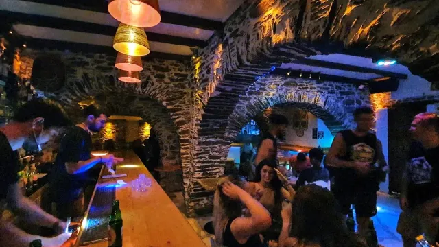 דלפק עץ ארוך וקהל צפוף בחלל אבן, עם מנורות תלויות בלילה