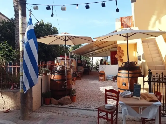 חצר עם שמשיות בהירות, שרשרת נורות תלויה, חביות עץ ודגל יווני על עמוד
