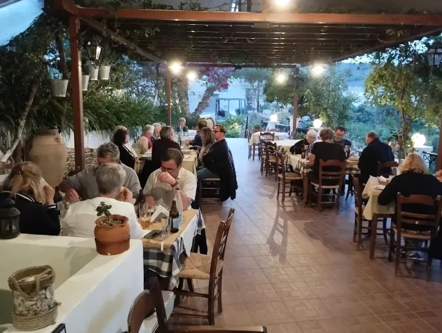 מרפסת מקורה בגפנים בערב, סועדים רבים ליד שולחנות לבנים ומנורות תלויות