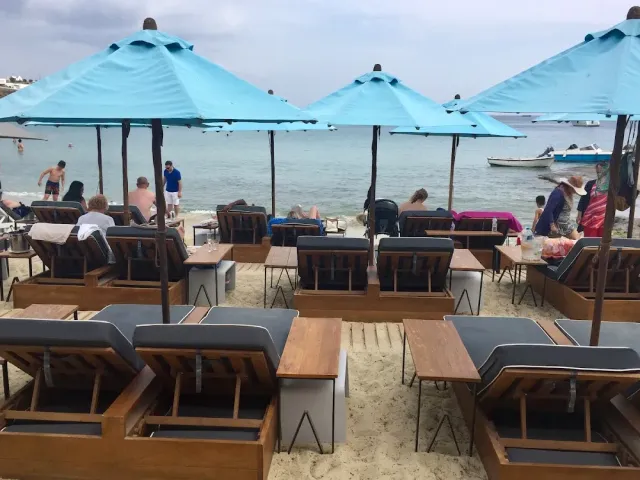 שמשיות כחולות ומיטות שיזוף מעץ על החול מול הים, עם מתרחצים ברקע