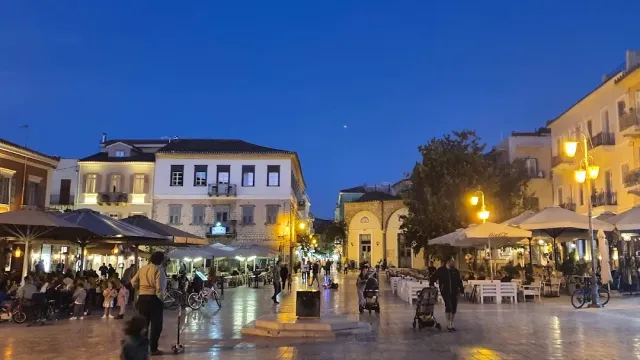 כיכר מרוצפת בשעת ערב, פנסים דולקים, שמשיות בתי קפה ובניינים מוארים