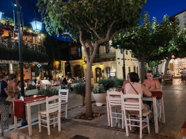 שולחנות לבנים בכיכר מרוצפת בערב, מתחת לעצים ופנסי רחוב דולקים