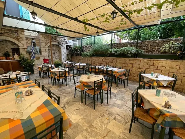 חצר מקורה עם מפות משובצות כתומות, כיסאות מתכת ירוקים, קיר אבן ופסל חזה