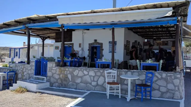 מבנה לבן עם סככת קנים וכיסאות כחולים, מעל קיר אבן